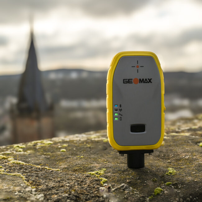 Eine GeoMax Zenith06 GNSS-Antenne auf einer steinige Mauer. Im Hintergund sieht man verschwommen auf Marburg herab.